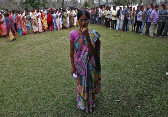 A woman shows her ink-marked finger after casting her vote as others line up to cast their ballot at a polling station in Nakhrai village in Tinsukia district
