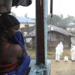 Liberian woman stands as health workers wearing protective clothing prepare to carry an abandoned dead body presenting with Ebola symptoms at Duwala market in Monrovia