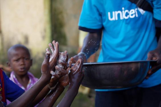 A social mobilizer from NGO (AJCOM), carries a basin of water past the soapy hands of children, to help prevent the spread Ebola in Conakry