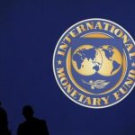 Visitors are silhouetted against the logo of the International Monetary Fund (IMF) in Tokyo