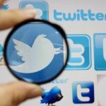 A person holds a magnifying glass over a computer screen displaying Twitter logos
