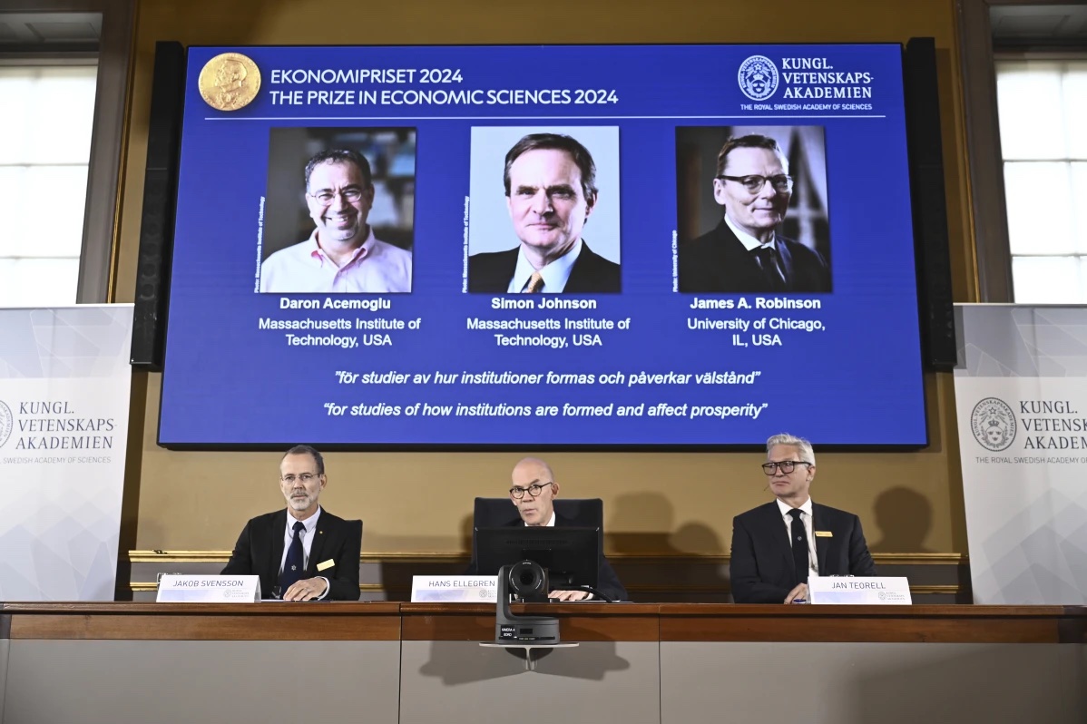 Academy of Sciences permanent secretary Hans Ellegren, center, Jakob Svensson, left, and Jan Teorell, of the Nobel assembly announce the Nobel memorial prize in economics winners, Daron Acemoglu, Simon Johnson and James A Robinson, seen on screen, during a press meeting at the Royal Swedish Academy of Sciences in Stockholm, Sweden, Monday Oct. 14, 2024.