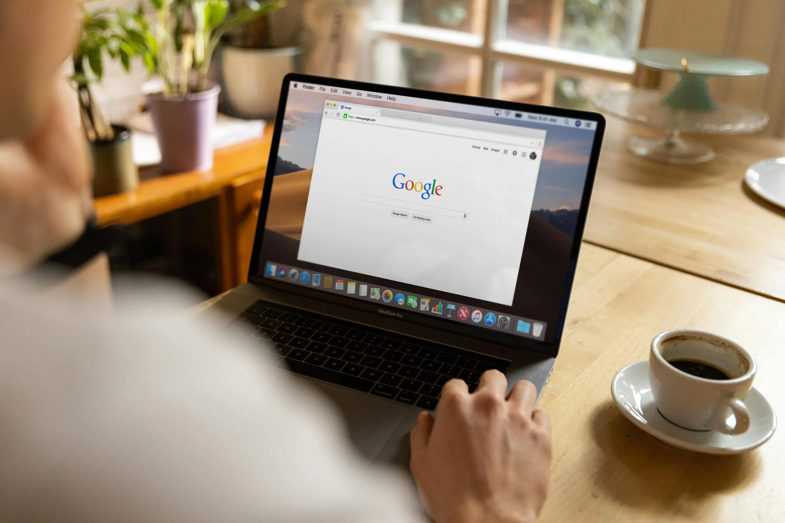 A man using a laptop, browsing Google homepage at a wooden table with coffee.
