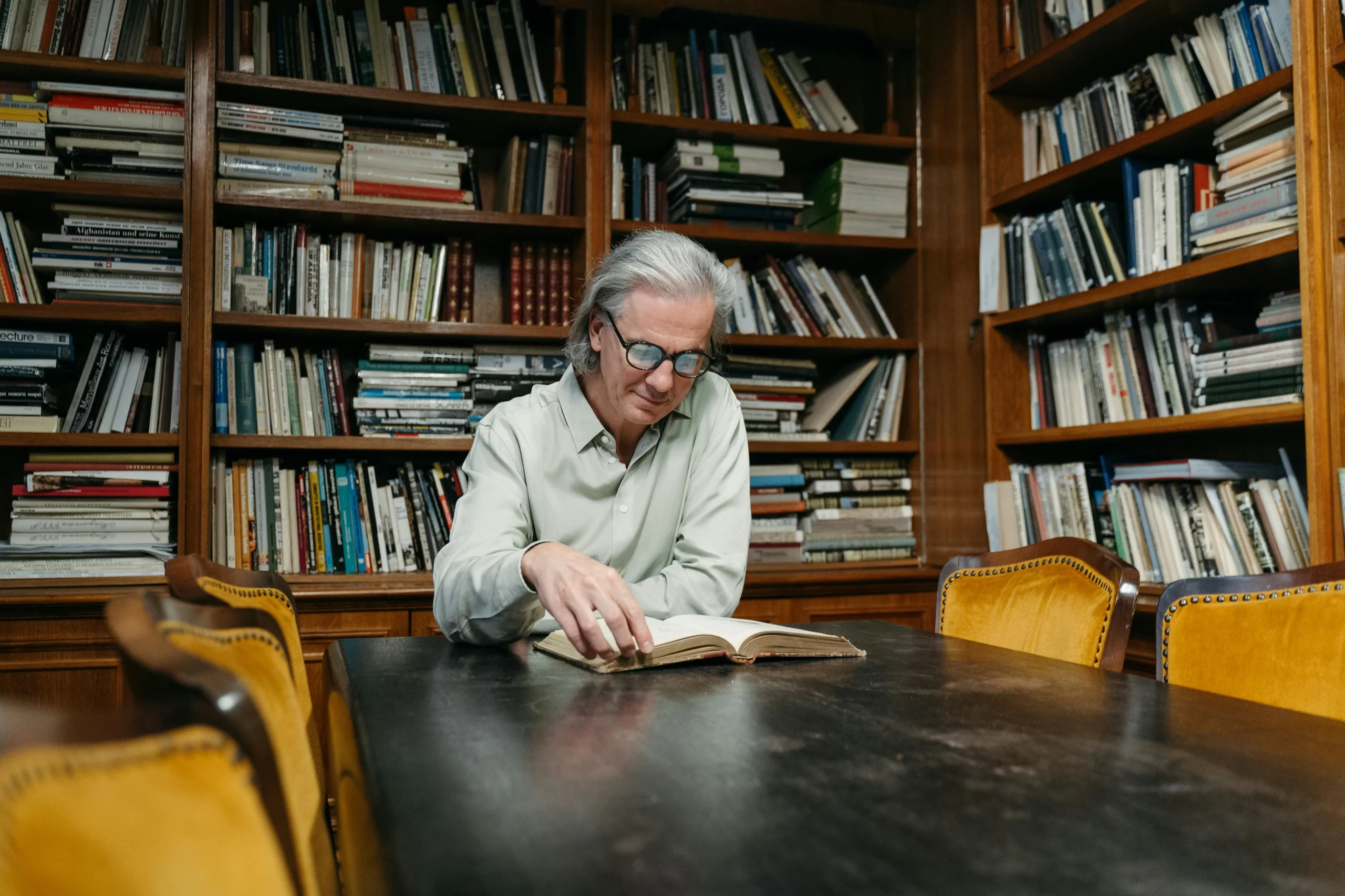 Senior man reading a book in a cozy library with wooden shelves filled with books. 