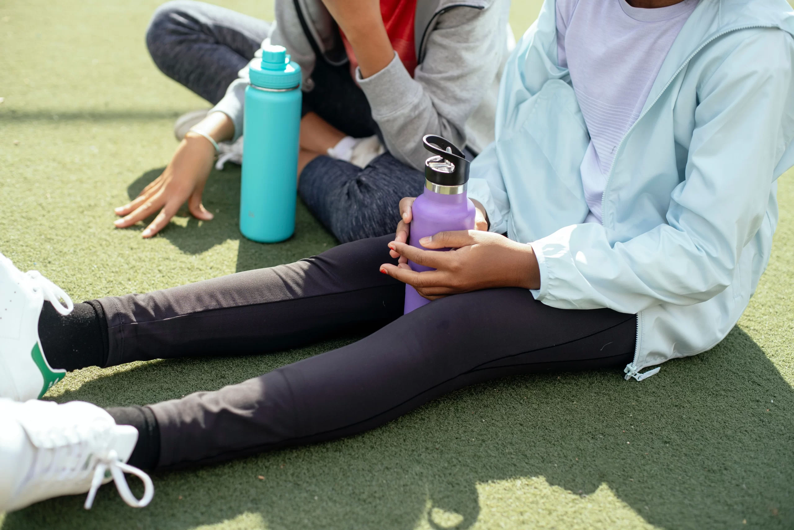 Two teenage sit on a sports field, holding reusable water bottles after an outdoor activity.