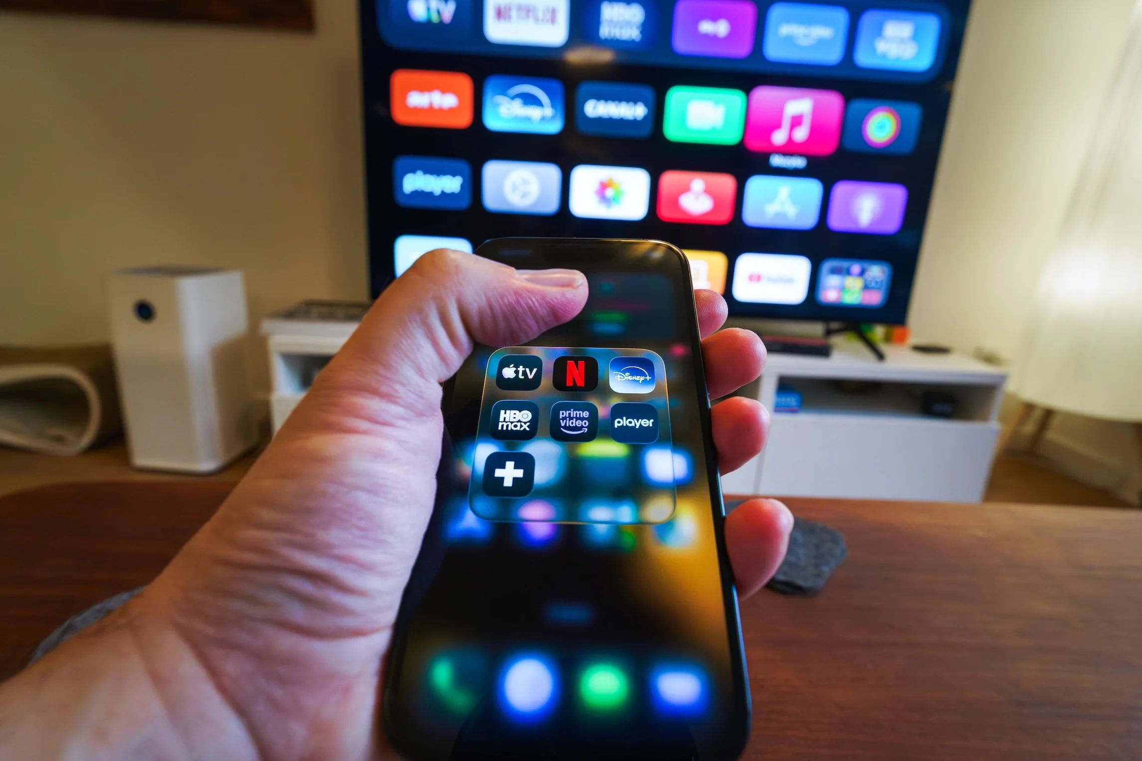 A hand holding a phone displaying streaming apps in front of a TV with multiple app icons.
